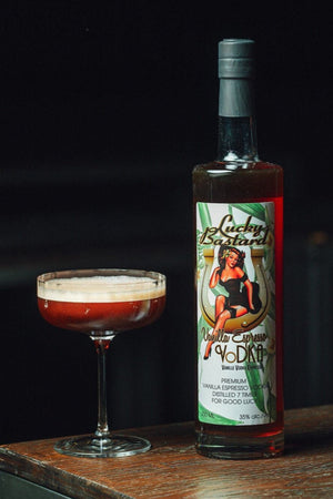 Bottle of Lucky Bastard Vodka on a wooden surface with a cocktail glass filled with a espresso martini