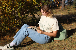 Person sitting on grass reading a book in a park with tennis & cocktails club t-shirt and blue jeans and new balance 550 sneakers