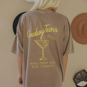 Person wearing a brown Cowboy Tears T-shirt with yellow text and graphics, standing in front of a wall with hats.