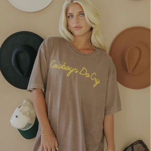 Woman wearing a brown t-shirt with 'Cowboys Do Cry' yellow text, standing in front of cowboy hats on a wall.