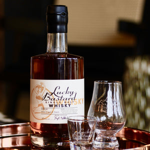 Bottle of Lucky Bastard Distillers SINGLE MALT WHISKY with two glasses on a copper tray. Made in Saskatoon Saskatchewan Canada with local ingredients. Barreled 2019.