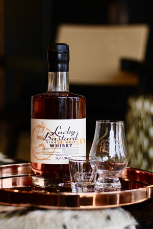 Bottle of Lucky Bastard Distillers SINGLE MALT WHISKY with two glasses on a copper tray. Made in Saskatoon Saskatchewan Canada with local ingredients. Barreled 2019.