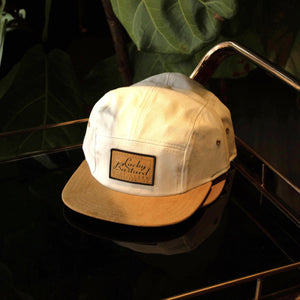 A beige and brown running hat with a label that reads 'Lucky Bastard'. The hat is displayed against a background with green foliage.