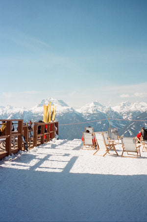 Snowy mountain top with wooden deck, chairs, and skiers against a clear blue sky.