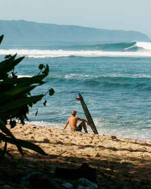 Person sitting on a beach with a surfboard, looking at the ocean waves.
