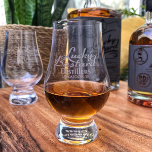 A clear glencairn glass labeled 'Lucky Bastard Distillers' filled with whisky, placed on a wooden surface with more glasses and a whisky bottle in the background.